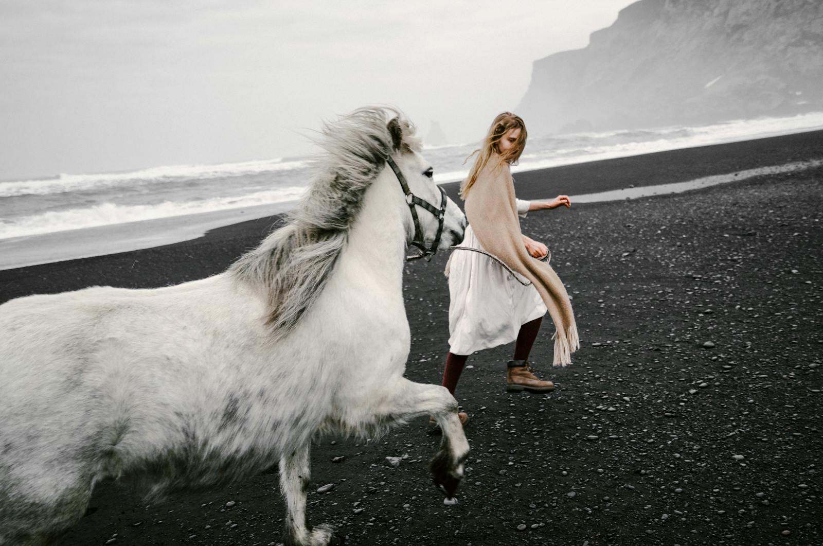 A young woman walks with a white horse on a rocky beach, capturing a scene of freedom and nature.